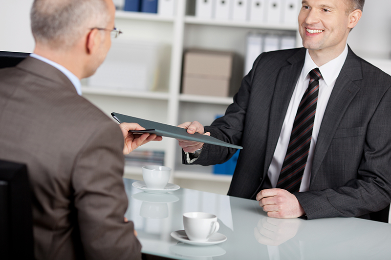 Smiling businessman hands client a folder over coffee.